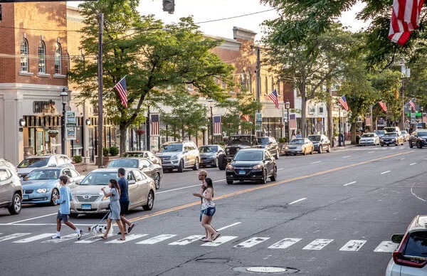 People walking across a street bordered by shops and cars